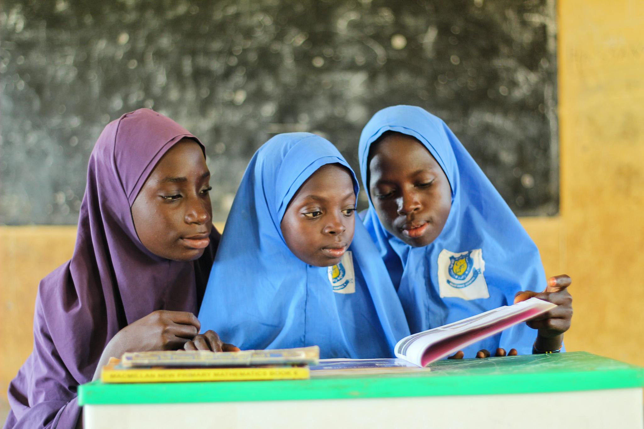 Three young Muslim girls in hijabs reading a book in a classroom setting, focused on learning.