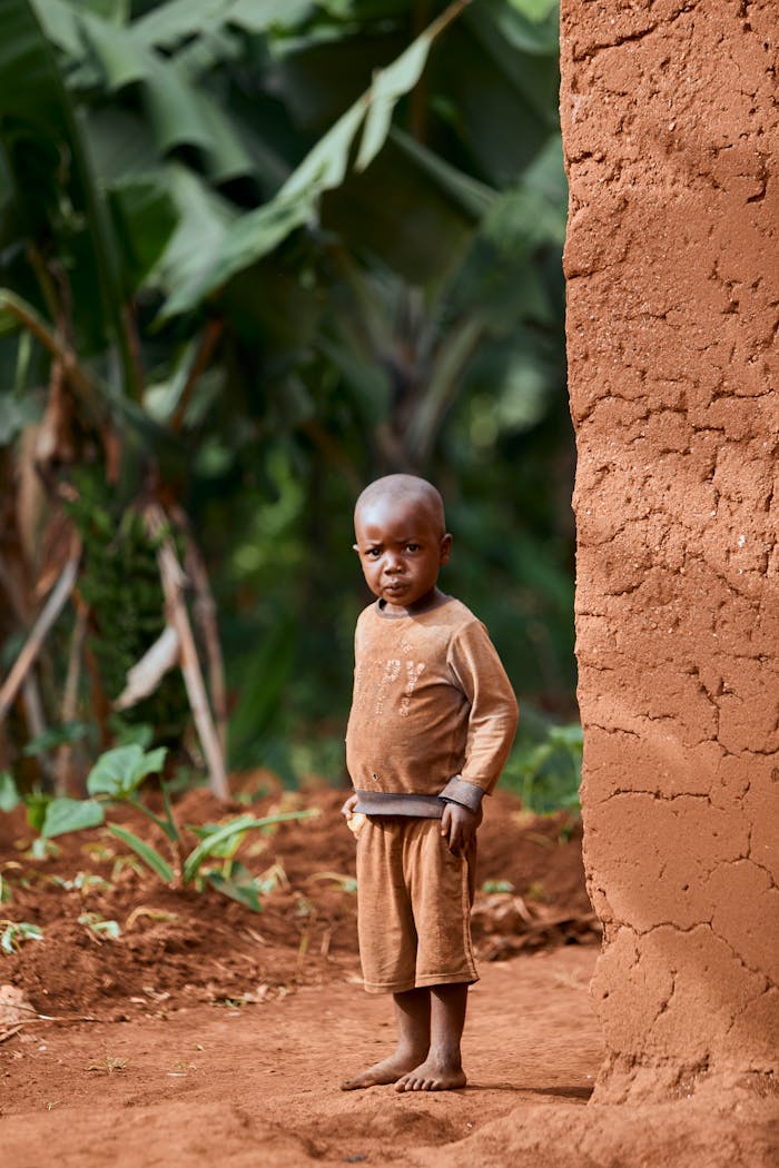 Young boy standing next to a clay wall with lush green foliage in the background.
