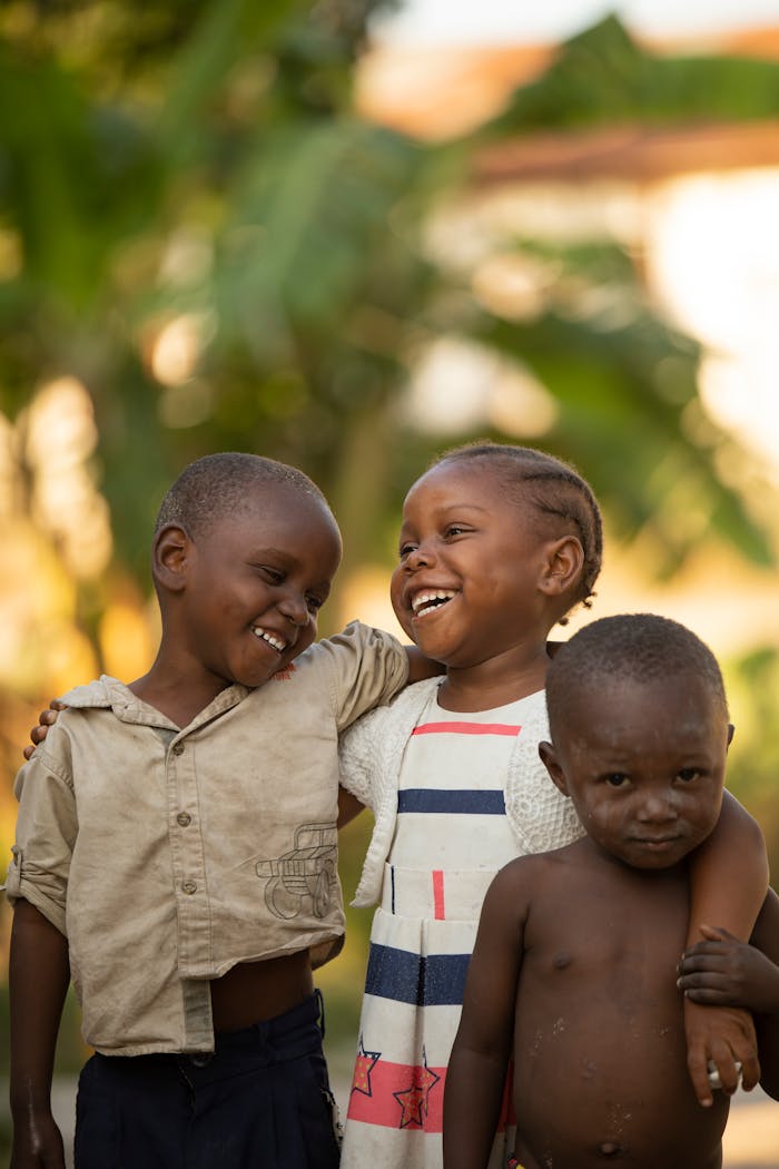 Laughing local African boys and girl in weathered clothes hugging and standing on street in sunny day