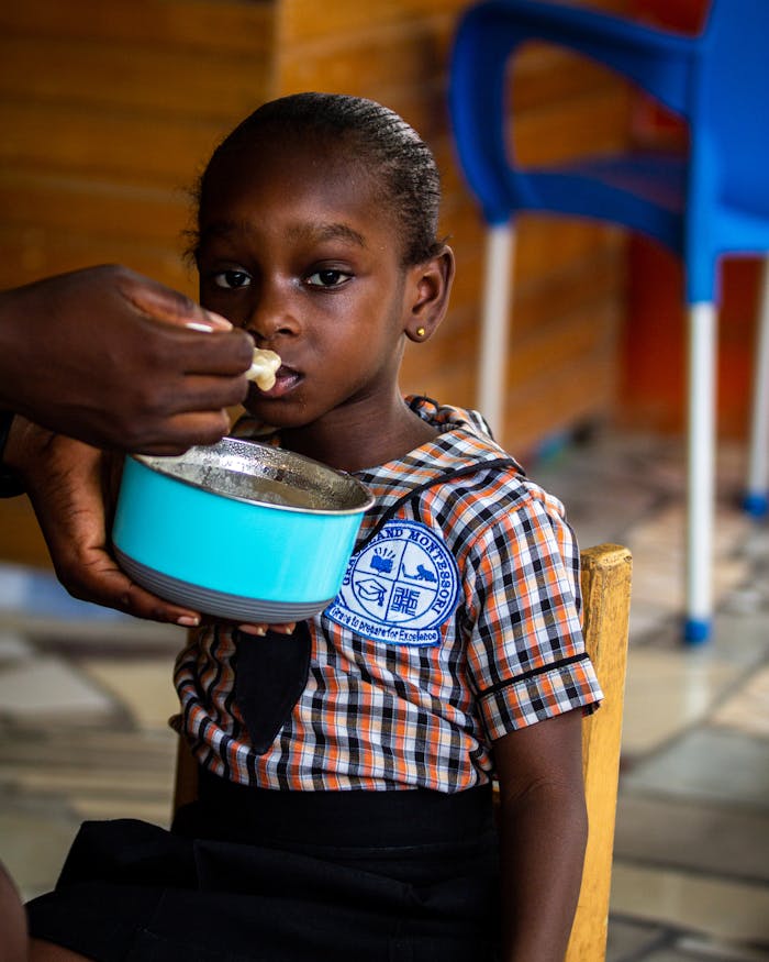 A young girl in a plaid uniform is being fed indoors, capturing a tender moment.